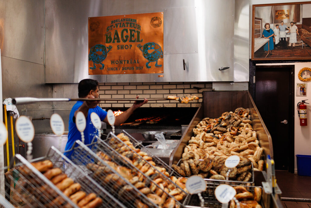 Worker dropping freshly made bagels from hot oven onto a large pile of bagels. 