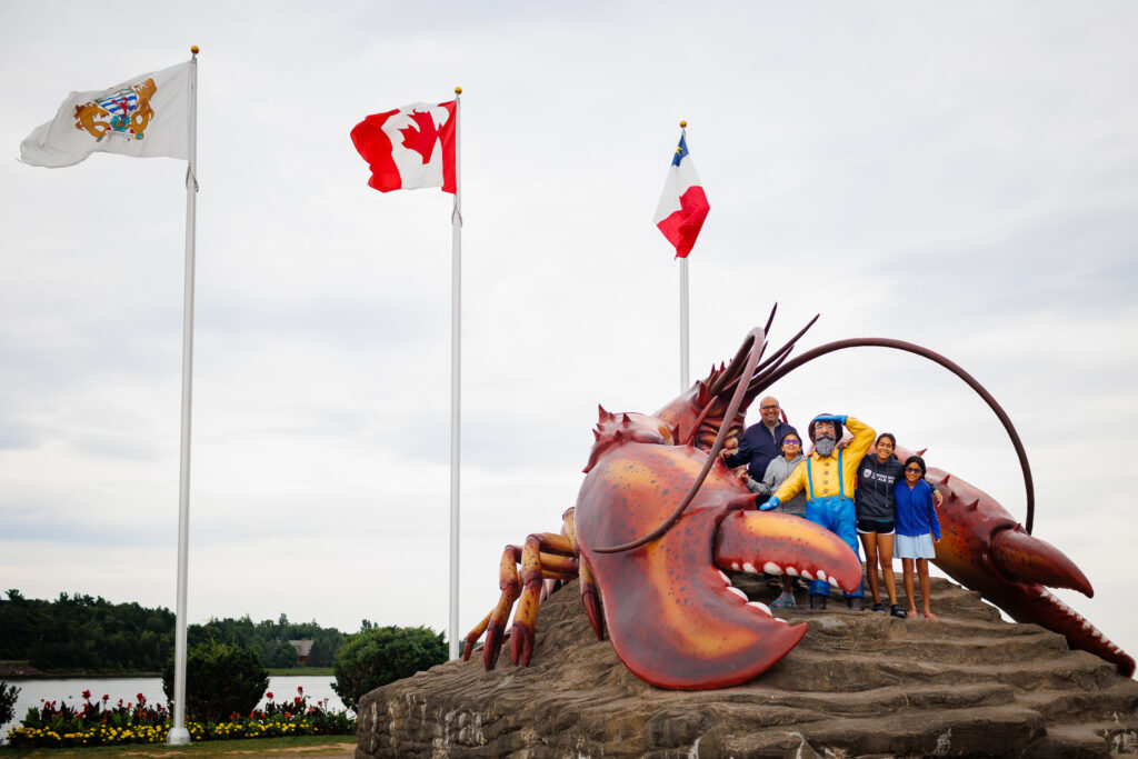 Family posing with fishermen statue in lobster statue with three flags waving in the wind.
