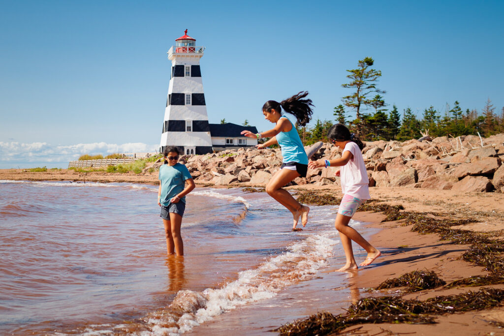 Three girls on red sand coastline with black and white striped lighthouse in background. One girl is jumping up to avoid the wave. 