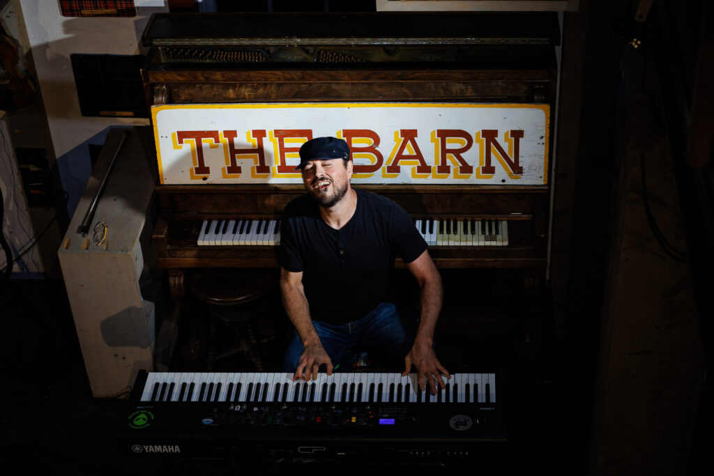 Pianist playing on a keyboard with a piano behind him with a "The Barn" sign. 