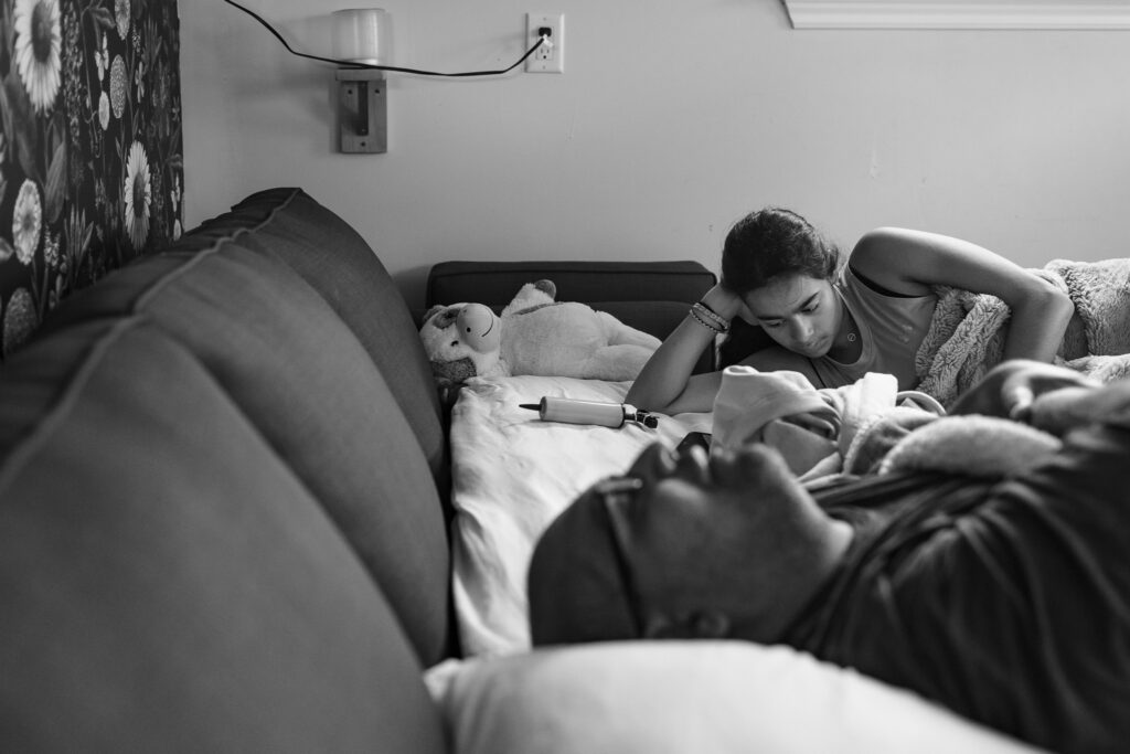 Girl reading while laying down with her father in glasses sleeping in the foreground. 