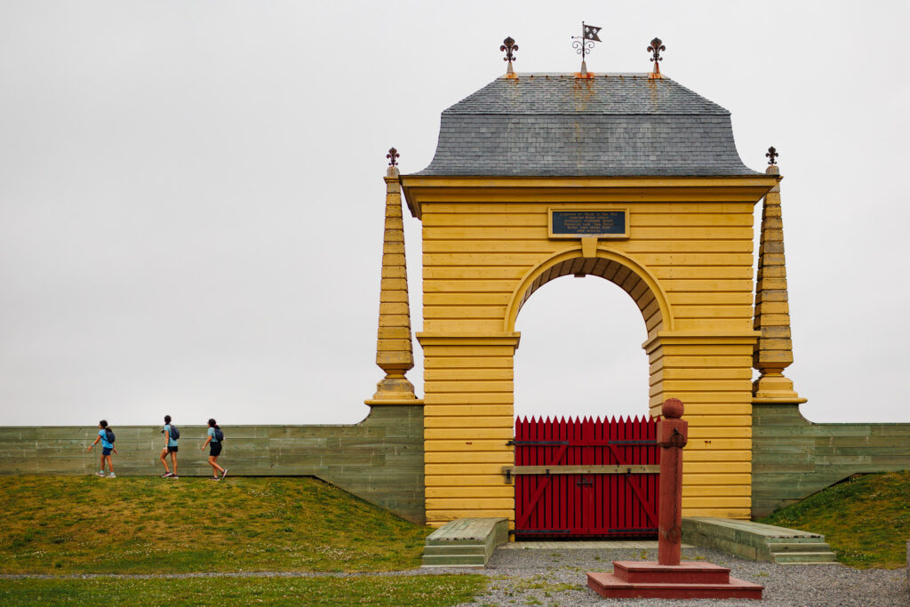 Three girls walking along the stone fence with a large archway and a red door. 