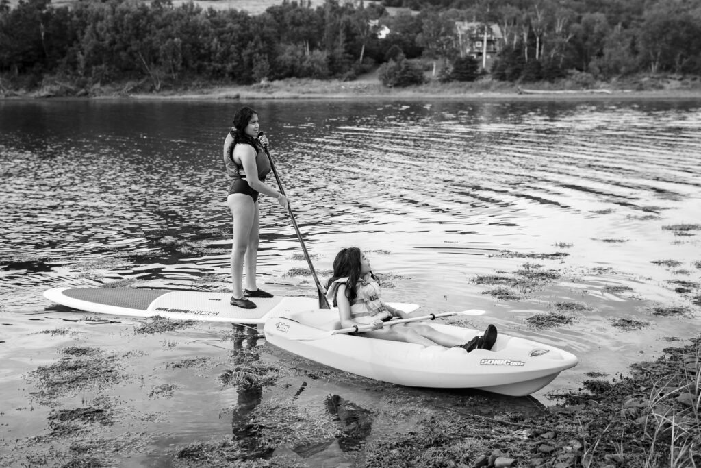 Girl on paddle board and girl in kayak coming up to the shore in lake in Nova Scotia. 
